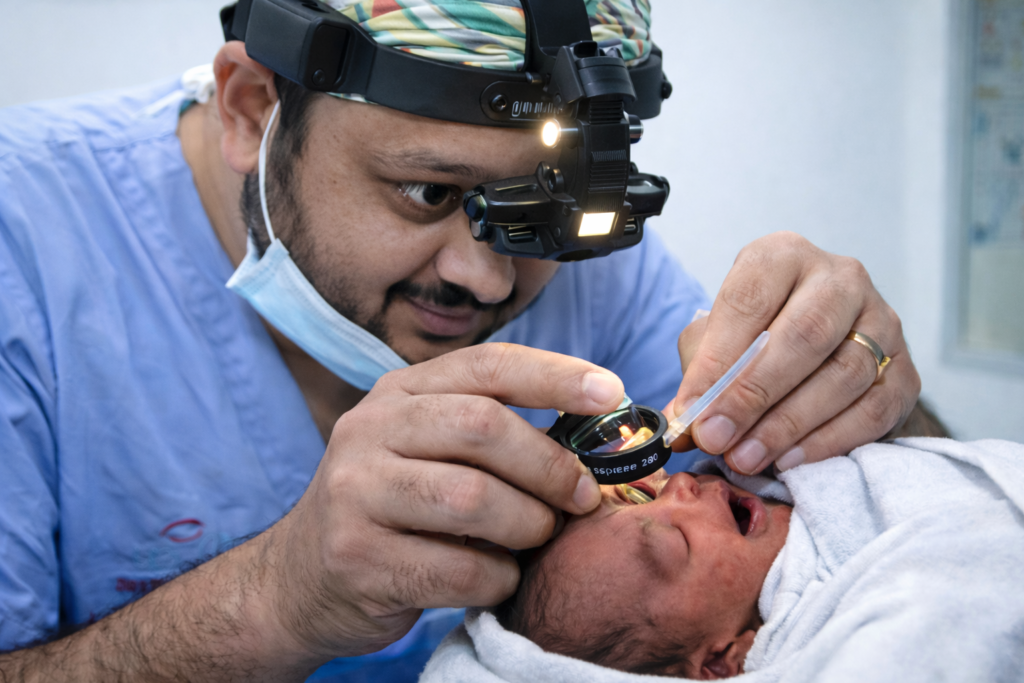 An ophthalmologist DR Jignesh Gala wearing surgical scrubs and an indirect ophthalmoscope examines a premature newborns eye for retinopathy of prematurity ROP using a handheld lens while the baby is swaddled in a white blanket in a clinical setting