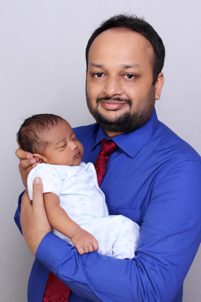 Dr Jignesh Gala in a blue shirt and red tie faces the camera while gently holding a sleeping newborn baby dressed in white against a plain background