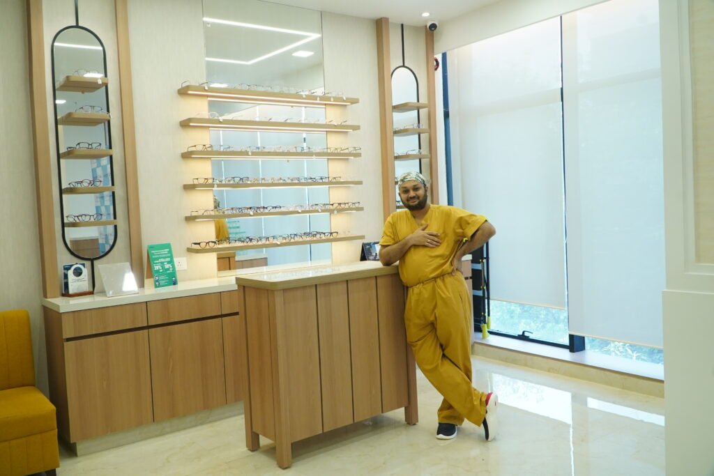 DrJignesh Gala retina specialist mumbai Crystal Clear Eye Clinic Best Eye Clinic in Andheri Mumbai A man in yellow scrubs leans against a wooden counter inside an eyewear store shelves of glasses behind him